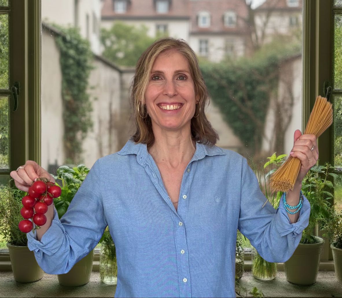 Debora Dumas lächelnd in ihrer Küche, mit frischen Rispentomaten und Spaghetti in den Händen
